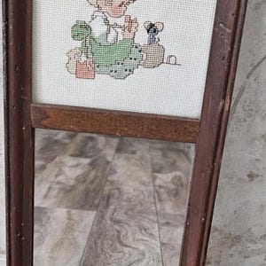 May include: A framed mirror with a cross-stitch design. The cross-stitch depicts a child in a green dress with a small animal. The frame is dark brown and rectangular.