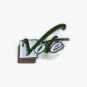 Puede incluir: Pin metálico con la palabra "Vote" en escritura blanca, acentuada con verde y una casilla de verificación blanca. Diseñado para ser usado como símbolo de compromiso cívico y participación política.