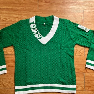 Puede incluir: Jersey de punto de cable verde con cuello en V blanco y ribetes blancos. El jersey tiene el número "1929" en la parte delantera.