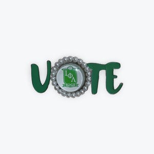 Può includere: Spilla in metallo verde che riporta la scritta "VOTE". La lettera "O" è un emblema circolare con sfondo bianco, stemma verde e bordo di perle. La spilla è progettata per essere indossata su abiti o accessori.