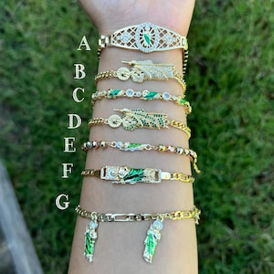 Könnte beinhalten: Sieben goldene Armbänder mit grünen und weißen Emaille-Anhängern. Die Anhänger zeigen religiöse Figuren und Symbole. Die Armbänder sind übereinander am Handgelenk einer Person getragen.