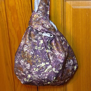 May include: A purple and gold fabric tote bag with a knotted handle. The bag has a marbled pattern with swirls of gold and purple.