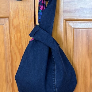 May include: A dark blue denim tote bag with a knotted handle. The handle features a floral pattern in pink, blue, and purple. The bag is hanging on a wooden door.