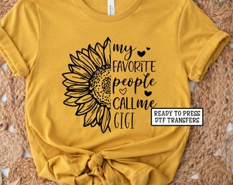 May include: A mustard yellow t-shirt with a black sunflower graphic and the text "my favorite people call me gigi" with hearts. The text "Ready to Press DTF Transfers" is in a white rectangle.