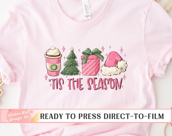 May include: Pink t-shirt with a festive graphic. The design includes a coffee cup, Christmas tree, gift, and Santa hat, with the text "'Tis the Season". The shirt is ready to press direct-to-film.