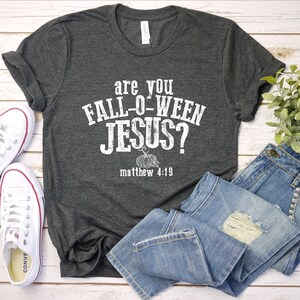 May include: Dark gray t-shirt with the text "are you FALL-O-WEEN JESUS?" and "matthew 4:19" in white. The shirt is paired with blue jeans and white sneakers. A small plant is also in the image.