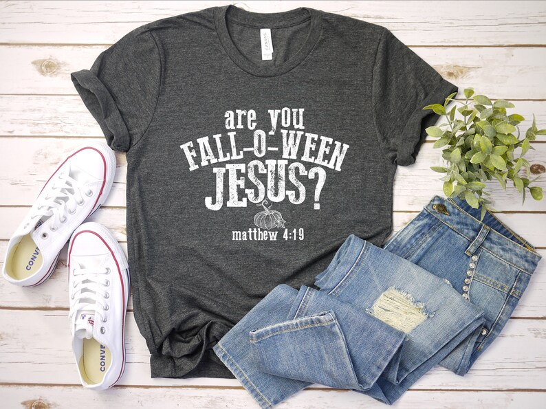 May include: Dark gray t-shirt with the text "are you FALL-O-WEEN JESUS?" and "matthew 4:19" in white. The shirt is paired with blue jeans and white sneakers. A small plant is also in the image.