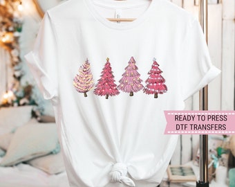 Puede incluir: Camiseta blanca con cuatro árboles de Navidad rosas con diferentes tonos y luces. Los árboles están dispuestos en fila, cada uno con un diseño único. El texto "READY TO PRESS DTF TRANSFERS" se muestra en una pancarta rosa.