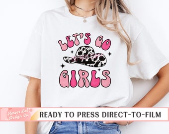 May include: White t-shirt with a pink and black graphic design that says "Let's Go Girls" with a cow print hat. The design is in a retro style.