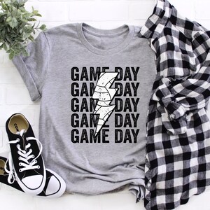 May include: A grey t-shirt with a white lightning bolt graphic and the text "GAME DAY" repeated five times. The shirt is laid flat on a white brick background.