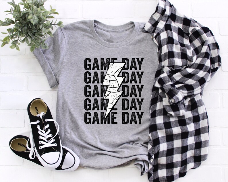 May include: A grey t-shirt with a white lightning bolt graphic and the text "GAME DAY" repeated five times. The shirt is laid flat on a white brick background.