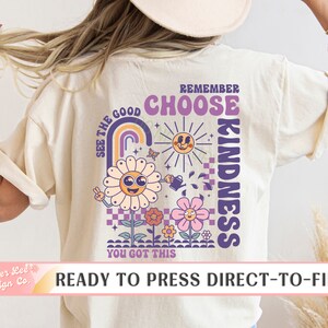 May include: A beige t-shirt with a purple and yellow graphic design featuring a flower, a rainbow, a sun, and the text "See the good, Remember Choose Kindness, You got this".