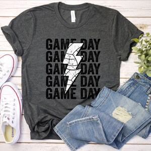 May include: A dark grey t-shirt with a white lightning bolt graphic and the text "GAME DAY" repeated five times. The shirt is laid flat on a wooden surface with a pair of white trainers and a pair of blue jeans.