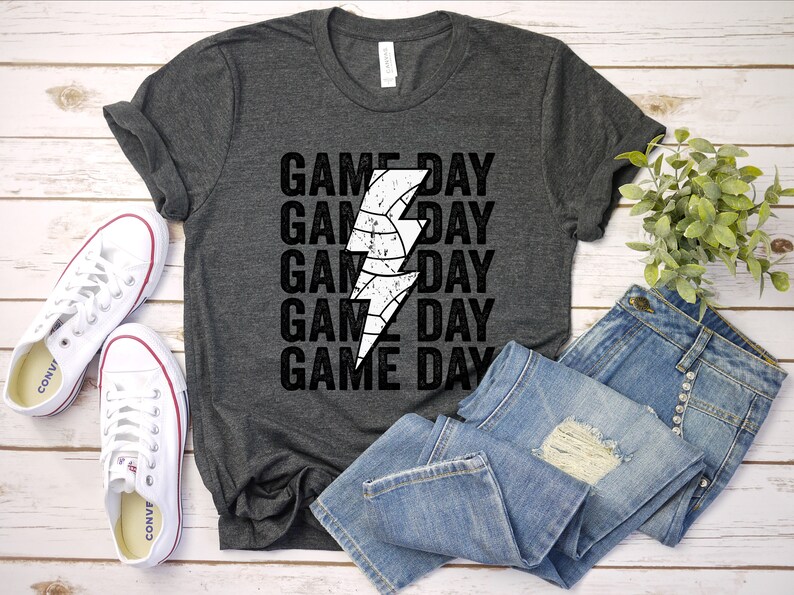 May include: A dark grey t-shirt with a white lightning bolt graphic and the text "GAME DAY" repeated five times. The shirt is laid flat on a wooden surface with a pair of white trainers and a pair of blue jeans.