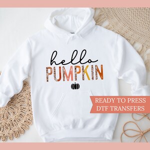 May include: White hoodie with a front pocket, featuring the text "hello PUMPKIN" in black and orange lettering, with a small black pumpkin graphic. The words "READY TO PRESS DTF TRANSFERS" are also visible.