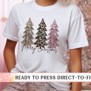 May include: White t-shirt featuring three sequined Christmas trees in gold, brown, and pink. The words "Merry Christmas" are written in a cursive font below the trees. The shirt is a ready-to-press direct-to-film design.