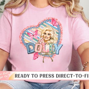May include: Pink t-shirt featuring a heart-shaped design with a portrait of a woman, butterflies, and the name "DOLLY" in a colorful font. The shirt has short sleeves and a crew neck.