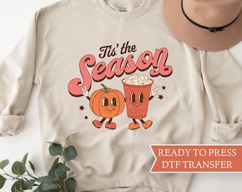 May include: Beige sweatshirt with the text "Tis' the Season" in a retro font. The design features a cartoon pumpkin and a coffee cup, both with smiling faces. A brown hat is placed on the top right.