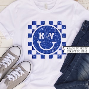 May include: White t-shirt with a blue and white checkered smiley face design featuring the letters "KY" inside the smiley face. The text "READY TO PRESS DIRECT TO FILM" is on a white banner.