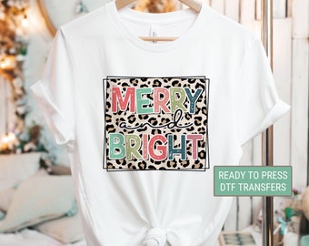May include: White t-shirt with the words "MERRY and BRIGHT" in colorful letters on a leopard print background. The shirt is hanging on a wooden hanger. A small green rectangle reads "READY TO PRESS DTF TRANSFERS".