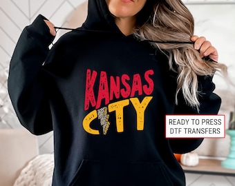 May include: Black hoodie with a red and yellow graphic that says "Kansas City" with a lightning bolt in the middle. The graphic has a distressed look. The text "READY TO PRESS DTF TRANSFERS" is in red at the bottom right corner of the image.