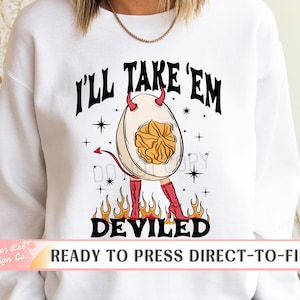 May include: White sweatshirt with a graphic of a deviled egg wearing red boots and devil horns, with the text "I'LL TAKE 'EM DEVILED". The design includes flames and stars. The bottom of the image reads "READY TO PRESS DIRECT-TO-FILM".