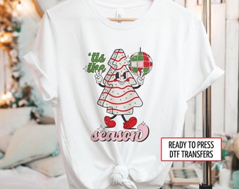 Puede incluir: Camiseta blanca con un personaje de árbol de Navidad de dibujos animados sosteniendo un signo de la paz y una bola de discoteca. El texto dice "'tis the season". El árbol tiene rayas rojas y zapatos rojos. La camiseta es una transferencia DTF.