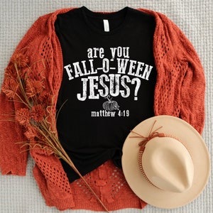 May include: A black t-shirt with the text "are you FALL-O-WEEN JESUS?" and a pumpkin graphic. The shirt is paired with a rust-colored cardigan, a beige hat, and decorative fall foliage. The ensemble is arranged on a neutral-toned surface.