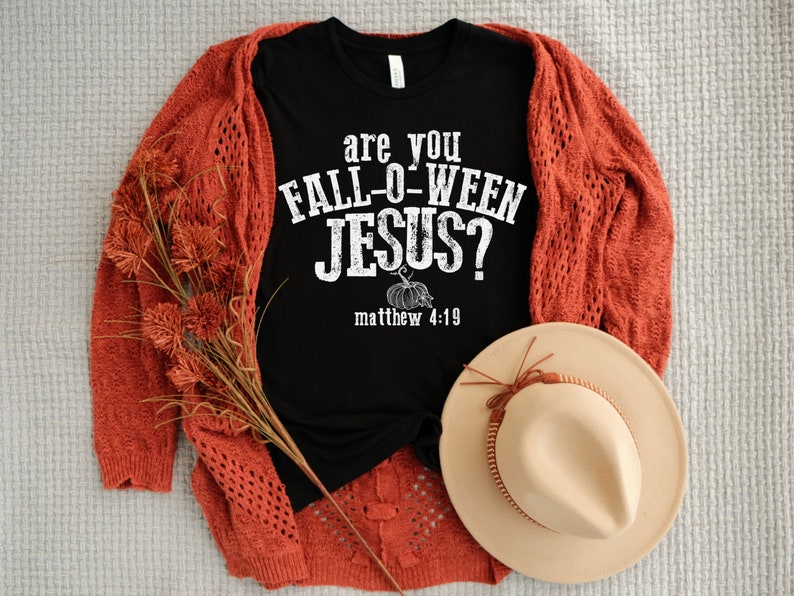 May include: A black t-shirt with the text "are you FALL-O-WEEN JESUS?" and a pumpkin graphic. The shirt is paired with a rust-colored cardigan, a beige hat, and decorative fall foliage. The ensemble is arranged on a neutral-toned surface.
