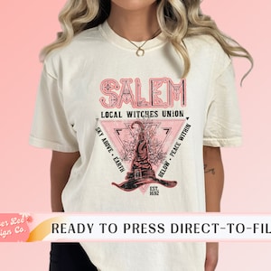 May include: Cream-colored t-shirt featuring a pink and black graphic with the text "SALEM LOCAL WITCHES UNION." The design includes a witch's hat, a triangle, and the words "SKY ABOVE EARTH" and "BELOW PEACE WITHIN." The text "EST. 1692" is also present.