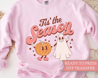May include: A light pink sweatshirt with a retro design featuring a smiling pumpkin and ghost holding hands. The text "Tis' the Season" is in a vintage font. The words "READY TO PRESS DTF TRANSFER" are also visible.