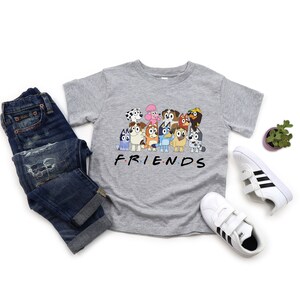 May include: Grey t-shirt featuring a graphic of the characters from the show "PAW Patrol" with the word "FRIENDS" printed below. The shirt is styled with blue jeans and white trainers.