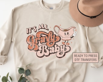 May include: Beige sweatshirt with the phrase "It's All Gravy Baby" in retro lettering. A cartoon gravy boat with a peace sign is featured. The design is in shades of brown, orange, and pink. A tan hat is in the background.