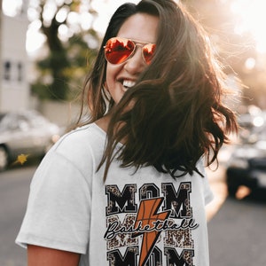 May include: White t-shirt with a graphic design featuring the words "MOM Basketball MOM" in black and white with a leopard print background. A lightning bolt and basketball are in the center of the design.