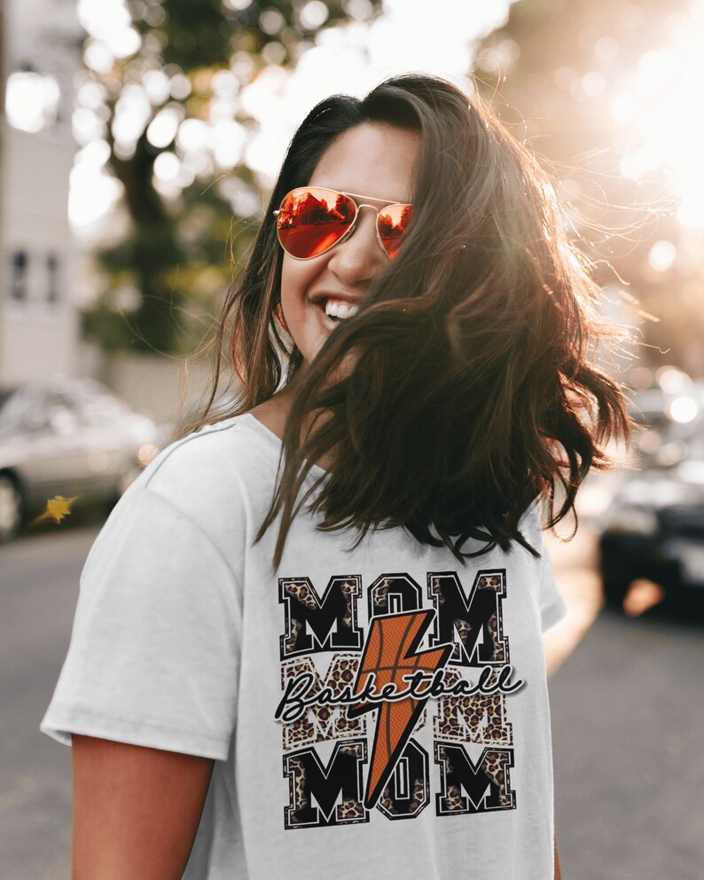 May include: White t-shirt with a graphic design featuring the words "MOM Basketball MOM" in black and white with a leopard print background. A lightning bolt and basketball are in the center of the design.