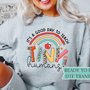 May include: A gray sweatshirt with the text "It's a good day to teach tiny humans" in colorful letters with a rainbow and a red apple. The text is surrounded by school supplies like a pencil, ruler, and a book. The sweatshirt is labeled "Ready to Press DTF Transfers".