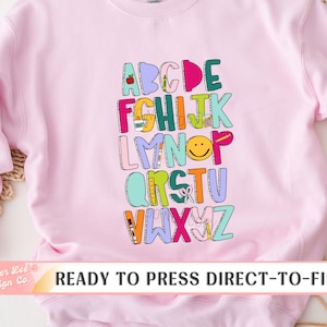 May include: A pink sweatshirt with a colorful alphabet design featuring school supplies and a smiley face. The text "READY TO PRESS DIRECT-TO-FILM" is at the bottom of the image.