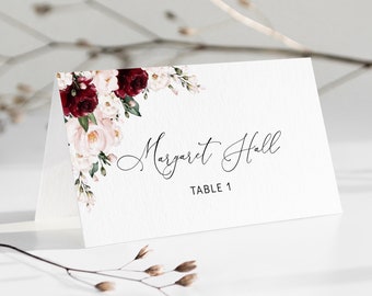 May include: A white place card with a floral design in shades of burgundy and pink. The text "Margaret Hall" and "Table 1" is printed in black.