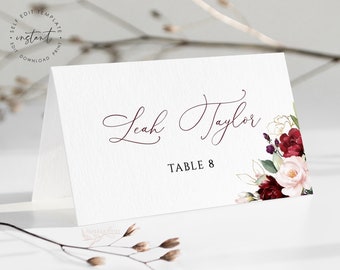May include: A white place card with burgundy floral accents and gold foil. The text reads "Leah Taylor" and "Table 8".