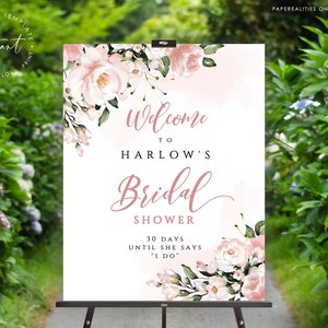 Op de afbeelding: Een wit welkomsbord voor een bruiloft met roze aquarelbloemen en de tekst "Welkom op de bruiloft van Harlow 30 dagen tot ze "Ja" zegt"