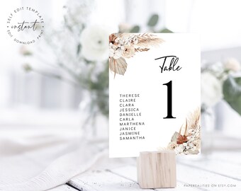 Puede incluir: Una tarjeta blanca con un diseño floral y el texto "Mesa 1" impreso en ella. La tarjeta está sentada en un soporte de madera. Los nombres de los invitados están listados debajo del número de mesa.