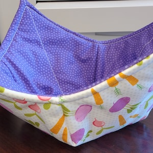 May include: A purple and white fabric bowl with a vegetable print. The bowl is made of two layers of fabric, with the purple layer on the inside and the white layer on the outside. The white layer has a print of carrots, turnips, and leaves.