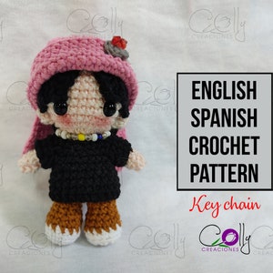 May include: A handmade crochet doll with a pink hat, black hair, and a black shirt. The doll has a beaded necklace and brown and white boots. The image includes text that says "English Spanish Crochet Pattern Key chain".