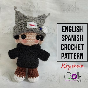 Könnte beinhalten: Häkelanleitung für einen grauen und braunen Amigurumi-Schlüsselanhänger. Der Schlüsselanhänger ist eine kleine Figur, die einen grauen Hut mit einem rot-weißen Patch und eine schwarze Jacke trägt. Die Anleitung ist in Englisch und Spanisch verfügbar.