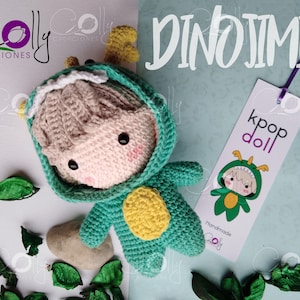 Puede incluir: Una muñeca de dinosaurio de crochet que lleva un disfraz de dinosaurio verde con detalles amarillos. La muñeca tiene el pelo castaño claro y la cara rosa. La muñeca está sentada sobre una superficie blanca con hojas verdes y una etiqueta que dice "kpop doll" y "Handmade".