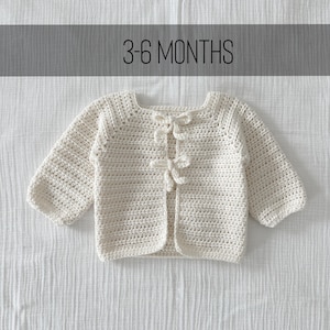May include: Cream-colored, crocheted baby cardigan with long sleeves and bow closures. The text "3-6 MONTHS" is displayed above the cardigan. The garment is made of a soft, textured yarn.