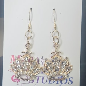 May include: A pair of gold-colored crown earrings with clear crystal accents. The earrings are dangling and have a post and hook closure. The earrings are displayed on a white card with the text "STUDIOS Fandom jewelry".
