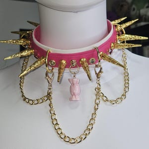 Könnte beinhalten: Ein leuchtend pinkes Halsband mit goldfarbenen Metallspitzen und runden Nieten. Das Halsband verfügt über zwei goldfarbene Ketten mit Karabinerverschlüssen und einen zentralen Anhänger in Form eines hellrosa verpackten Bonbons.