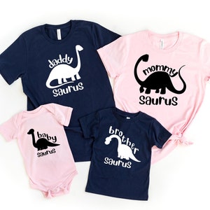 May include: A collection of matching family t-shirts and a baby onesie. The shirts are navy blue and pink, with white and black dinosaur graphics and the words "daddy saurus", "mommy saurus", and "brother saurus". The baby onesie is pink and says "baby saurus".