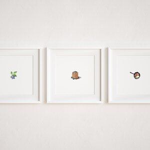 May include: Three white picture frames with white mats hanging on a white wall. The left frame shows a small sprig of blueberries. The middle frame shows a stack of pancakes with blueberries on top. The right frame shows a frying pan with a fried egg inside.